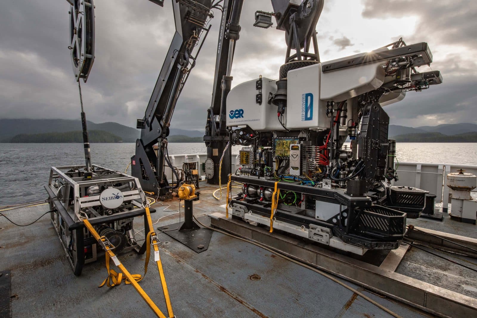 Two remotely operated vehicles secured on a ship's deck with a coastline behind them